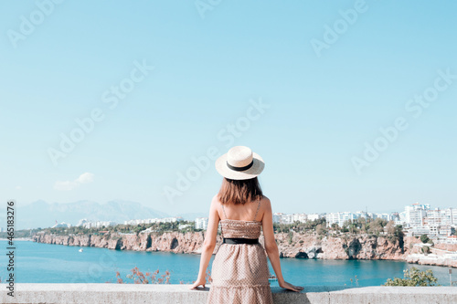 A young female tourist in a dress and hat on the viewpoint against the background of the city Antalya. Female tourist enjoying the cityscape © epovdima