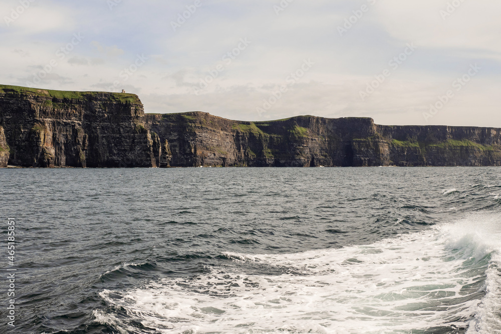 View on Cliff of Moher from a board of a ferry or cruise ship. West coast of Ireland. Popular tourist destination. National landmark. Cloudy sky. Calm surface of Atlantic ocean.