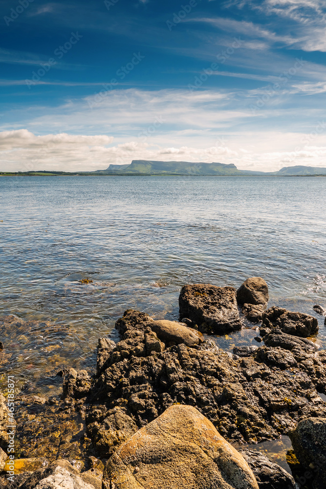Rough rocks by the ocean. Silhouette of Benbulben flat top mountain in the background. Beautiful blue cloudy sky. Warm sunny day. County Sligo, Ireland. Vertical image. Amazing nature scene