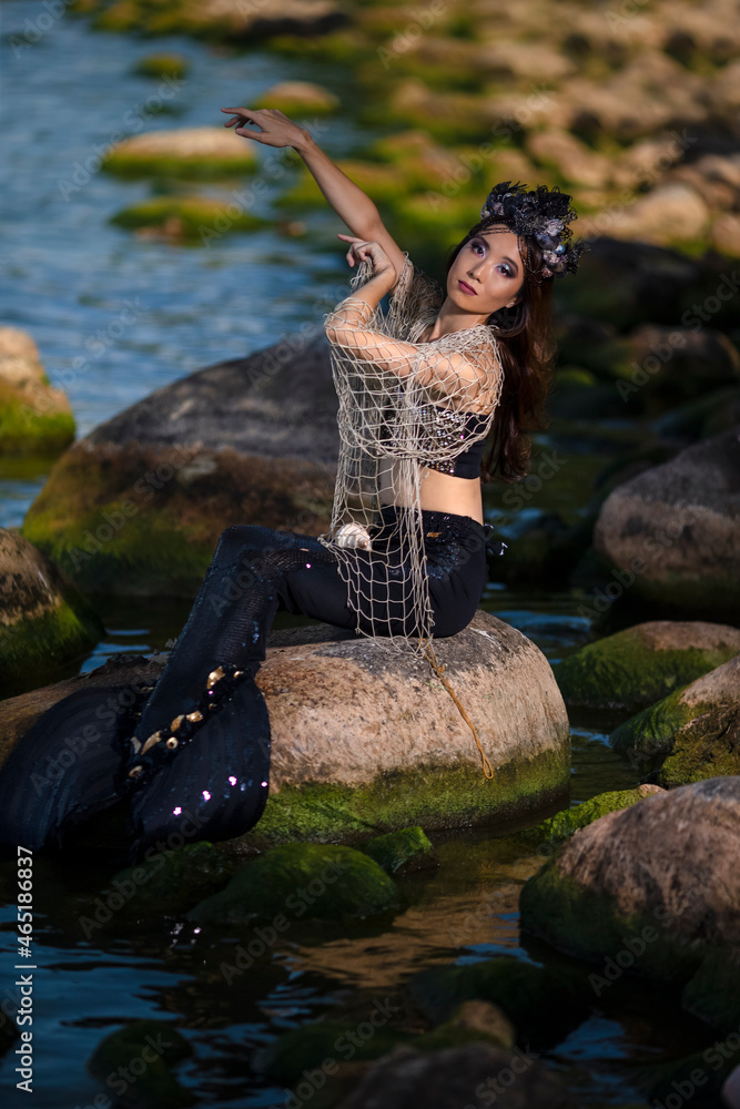 Asian Mermaid With Net At Sea Coast on Rocks While Wearing Seashell Decorated Crown and Black Shiny Tail On Sexy Body Covered With Strasses As Mistress Queen of Sea.