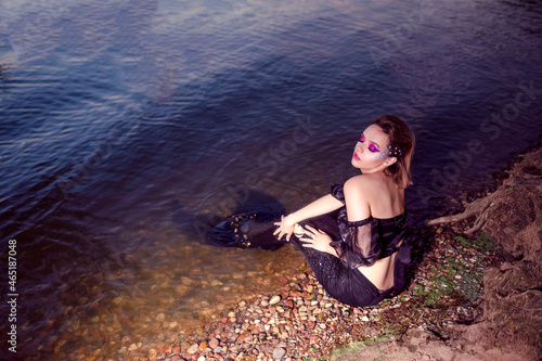 Fototapeta Naklejka Na Ścianę i Meble -  Mermaid Posing Near Sea Shore on Rocks While Wearing Artistic Makeup with facial Strassses and Black Shiny Tail Covered With Colorful Spangles Posing as Mistress Queen of Sea.
