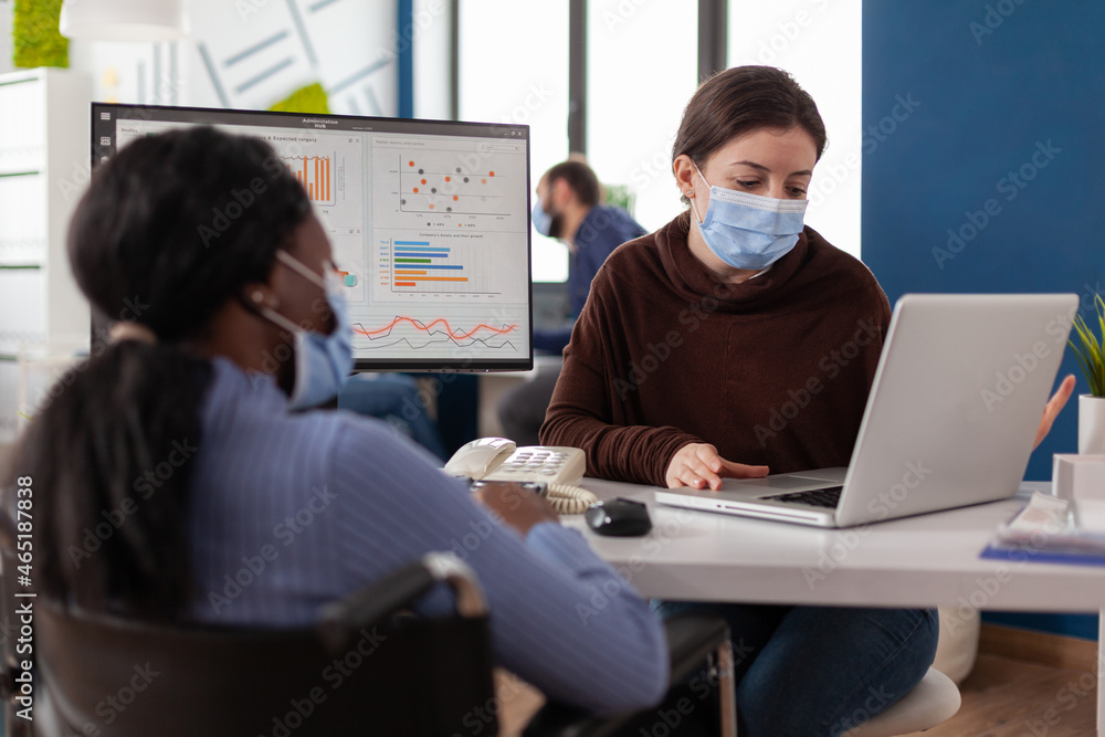 Multiethnic business team with medical face masks to prevent infection with coronavirus analyzing financial graph discussing strategy. Disabled paralyzed businesswoman working in startup office