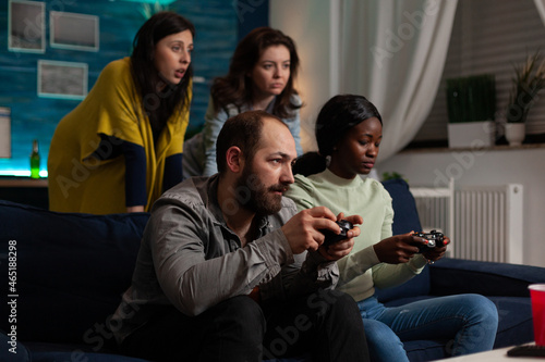 Serious concentrated multi-ethnic friends sitting on sofa holding joystick playing videogames during online competition enjoying fun wekeend at home. Group of people hanging out together