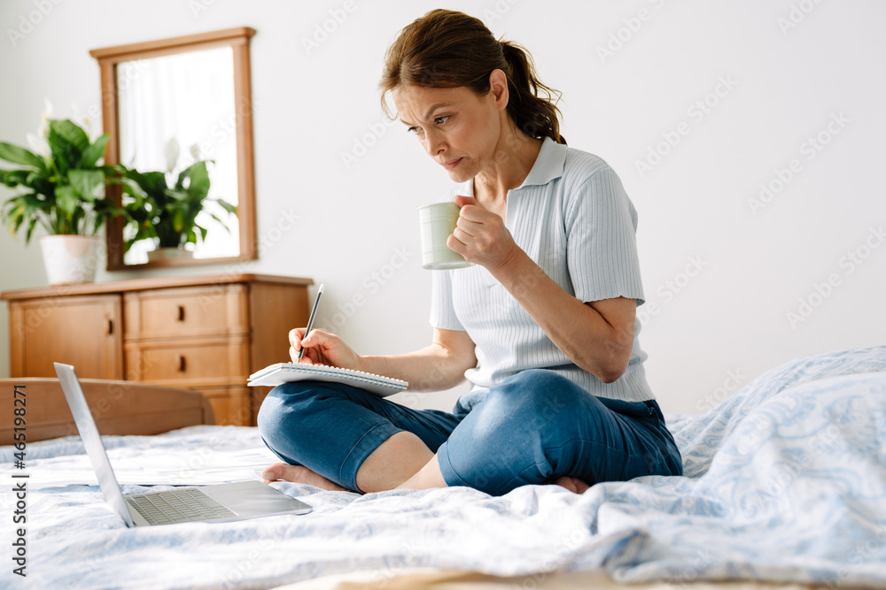 Ginger mature woman writing down notes while working with laptop at home Ginger mature woman writing down notes while working with laptop at home