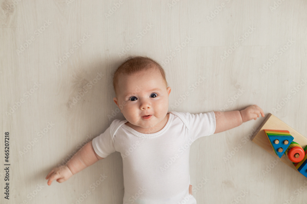 cheerful newborn baby in a white bodysuit lies on his back on the floor and plays with educational toys. products for children. concept of a happy childhood and motherhood. child care. space for text