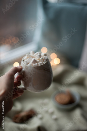 Hand holding a cup of coffee with marshmallows with christmas lights at cozy home
