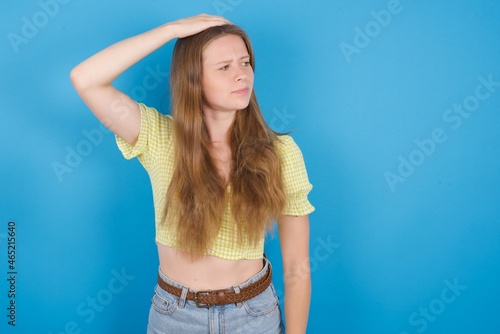 young ukranian girl wearing yellow t-shirt over blue backaground saying: Oops, what did I do? Holding hand on head with frightened and regret expression. photo