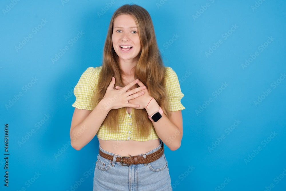 Happy smiling young ukranian girl wearing yellow t-shirt over blue backaground has hands on chest near heart. Human emotions, real feelings and facial expression concept. Happy smiling young ukranian girl wearing yellow t-shirt over blue backaground has hands on chest near heart. Human emotions, real feelings and facial expression concept.