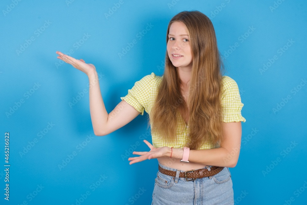 young ukranian girl wearing yellow t-shirt over blue backaground pointing aside with both hands showing something strange and saying: I don't know what is this. Advertisement concept. young ukranian girl wearing yellow t-shirt over blue backaground pointing aside with both hands showing something strange and saying: I don't know what is this. Advertisement concept.
