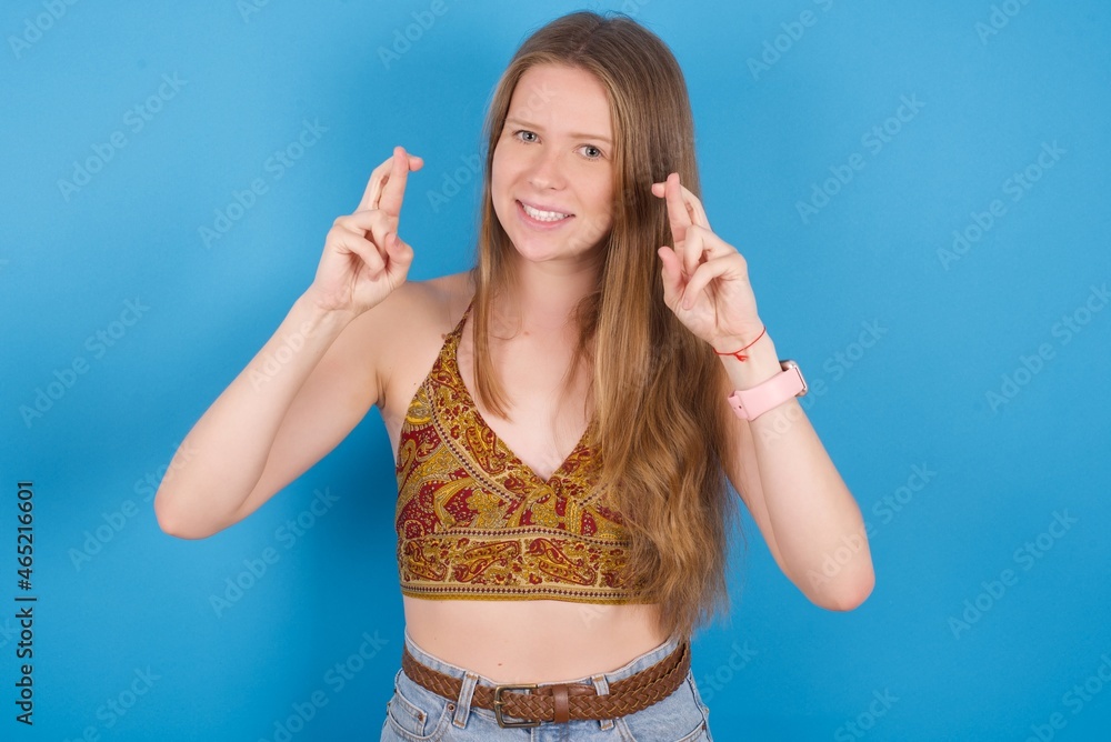 Fototapeta premium young ukranian girl wearing tank top over blue backaground holding fingers crossed with worried expression hoping boss didn't noticed mistakes at work.