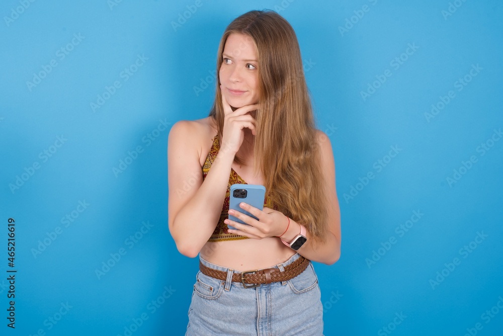 Thinking dreaming young ukranian girl wearing tank top over blue backaground using mobile phone and holding hand on face. Taking decisions and social media concept. Thinking dreaming young ukranian girl wearing tank top over blue backaground using mobile phone and holding hand on face. Taking decisions and social media concept.