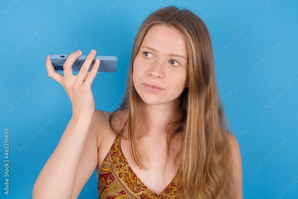 Smiling young ukranian girl wearing tank top over blue backaground listening a voice message from her smartphone. Communication and technology concept.