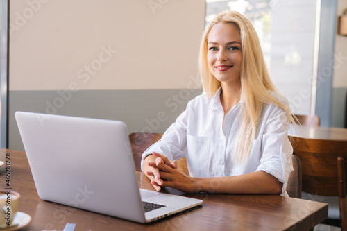 Medium shot portrait of young attractive smiling blonde woman sitting at table with laptop in cafe, looking at camera. Happpy beautiful businesswoman with laptop in coffee shop.
