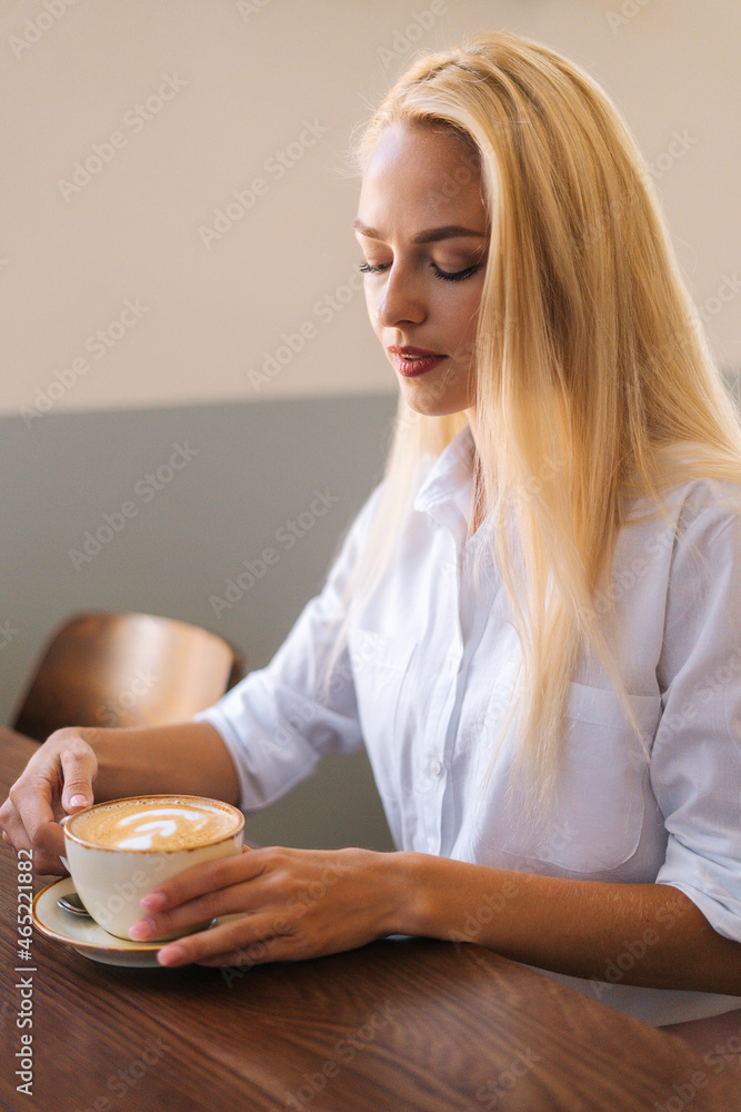 Side view of attractive young blonde woman holding cup of hot latte coffee with beautiful pattern sitting at table, looking down. Vertical portrait of cute female drinking coffee resting at cozy cafe. Side view of attractive young blonde woman holding cup of hot latte coffee with beautiful pattern sitting at table, looking down. Vertical portrait of cute female drinking coffee resting at cozy cafe.