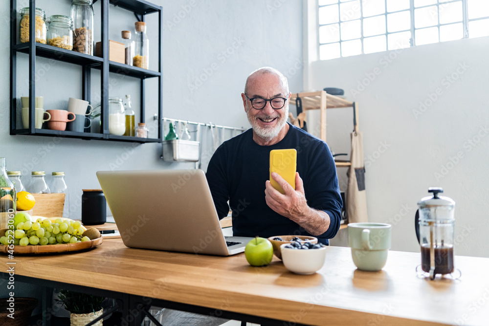 Smiling man with laptop using smart phone in kitchen Smiling man with laptop using smart phone in kitchen