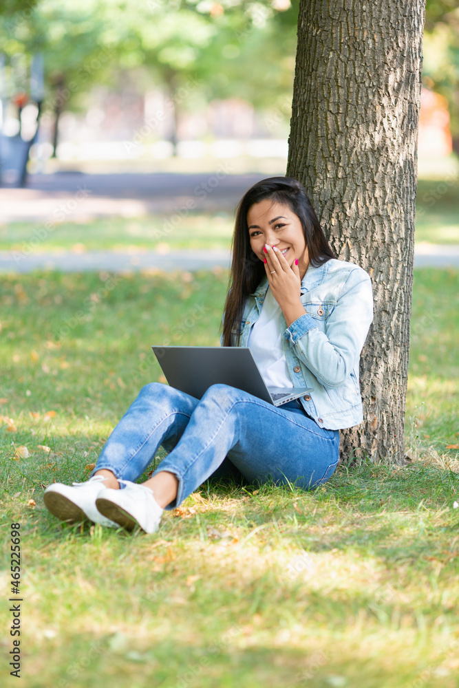 Vertical photo of an Asian young woman is sitting in the park on the grass with a laptop. Video call - she laughs, covering her mouth with her hand. Vertical photo of an Asian young woman is sitting in the park on the grass with a laptop. Video call - she laughs, covering her mouth with her hand.