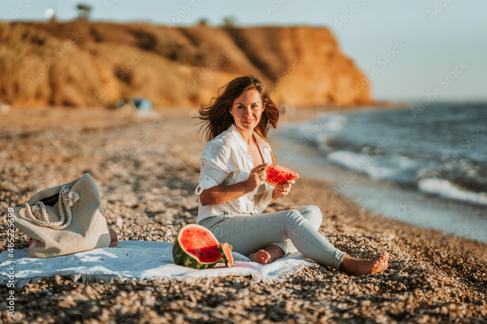 A young woman in a white shirt and jeans is sitting on the beach and eating a watermelon. The concept of a quiet summer evening and a picnic with fruit