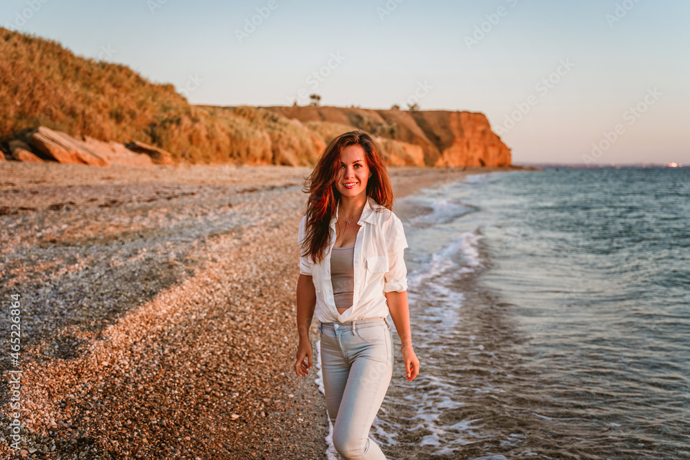 A charming young woman in a white shirt and jeans on a calm romantic beach by the sea. Summer woman vacations concept. The concept of active life.