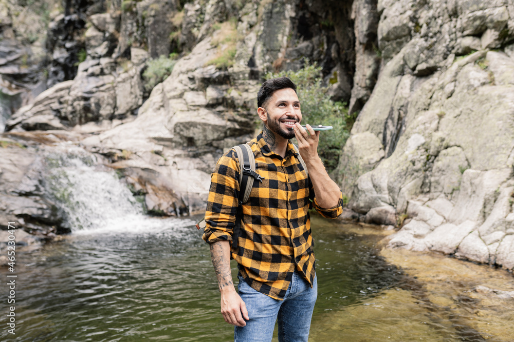 Smiling mid adult man talking on smart phone through speaker by pond Smiling mid adult man talking on smart phone through speaker by pond
