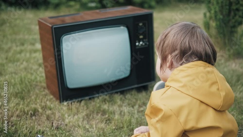 Small child is sitting on grass and watching an old retro TV. TV is strobing interference. Concept of post apocalypse, horror, surrealism and mysticism. Children dependence on TV, gadgets and devices photo