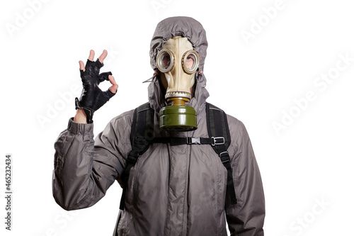 An isolated shot of a man dressed in a gas mask, a hooded jacket, a backpack, shows a gesture of ok, looks into the camera. On a white isolated background