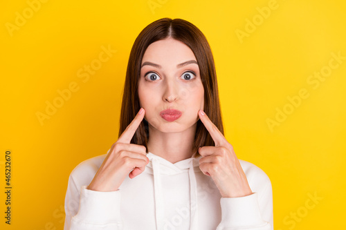 Photo of funny cute lady inflate cheeks press fingers wear white hoodie isolated yellow color background