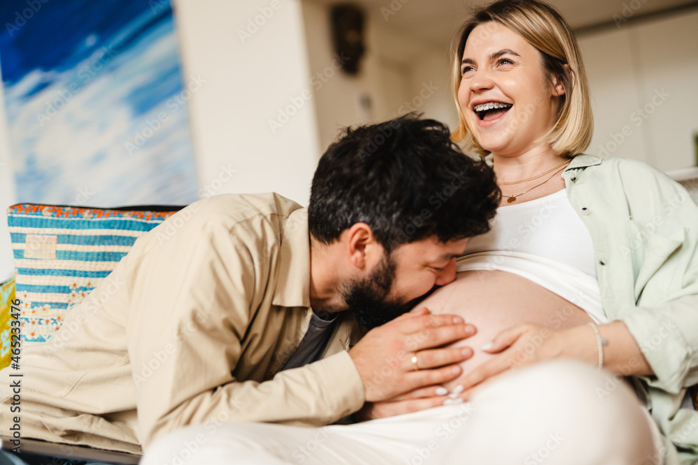 White man kissing his pregnant wife's belly while sitting on couch White man kissing his pregnant wife's belly while sitting on couch