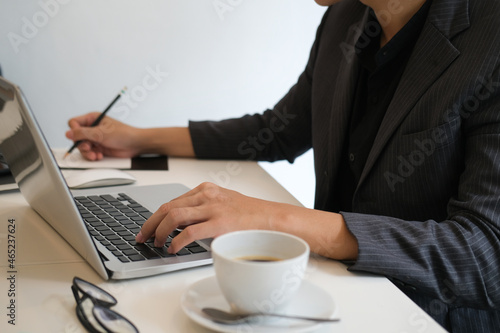 Cropped image of a businessman taking notes while typing on a computer laptop at the white working desk surrounded by a coffee cup and eyeglasses.