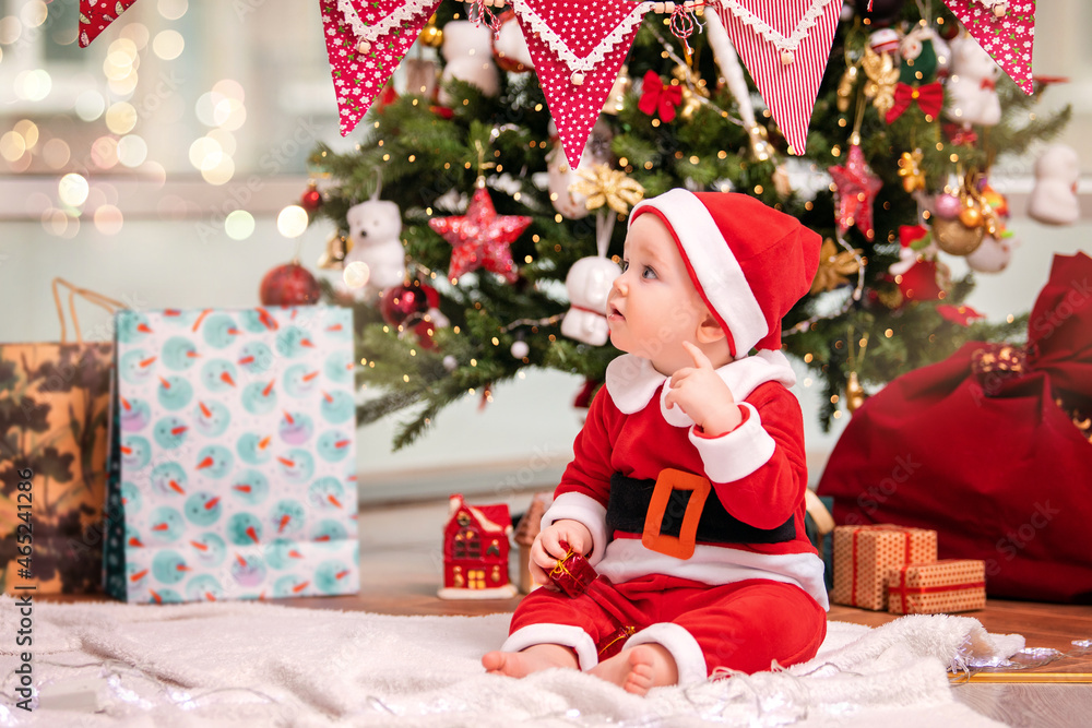 An adorable kid boy dressed as Santa Claus plays near a decorated Christmas tree in the living room.