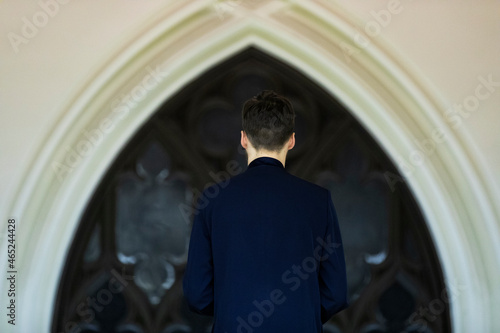 A young man dressed in blue standing in front of a church window contemplating about his faith