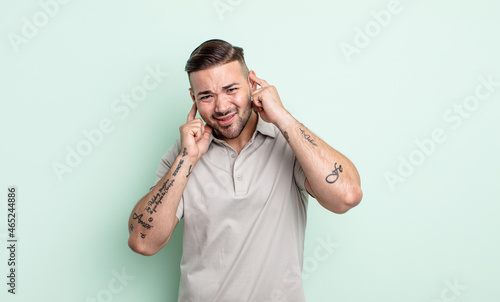 young handsome man looking angry, stressed and annoyed, covering both ears to a deafening noise, sound or loud music photo