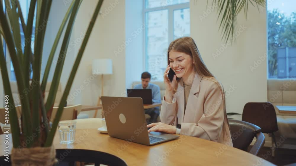 Young woman with long hair wearing jacket sitting in cafe, using laptop and talking happily on phone, man on blurred background working on laptop. Tracking shot of freelancer working remotely Young woman with long hair wearing jacket sitting in cafe, using laptop and talking happily on phone, man on blurred background working on laptop. Tracking shot of freelancer working remotely