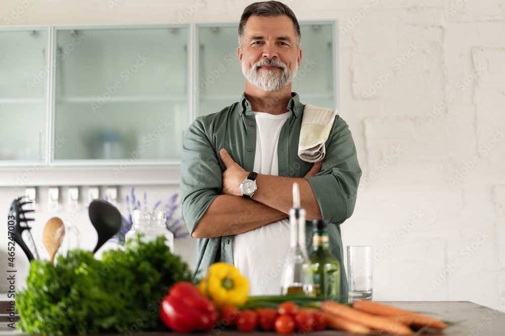 Smiling mature man standing in kitchen, looking at camera, making fresh vegetable salad enjoying cooking alone at home Smiling mature man standing in kitchen, looking at camera, making fresh vegetable salad enjoying cooking alone at home