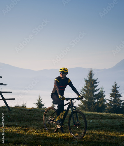 Fototapeta Naklejka Na Ścianę i Meble -  Man in cycling suit riding bicycle on grassy hill in the morning. Male bicyclist enjoying the view of majestic mountains during bicycle ride. Concept of sport, bicycling and nature.