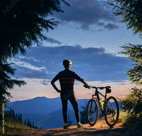 Fototapeta Naklejka Na Ścianę i Meble -  Rear view on a cyclist standing holding his bike, watching beautiful landscape, mountains peaks and sunset, surrounded with spruces. Square picture. Active lifestyle concept