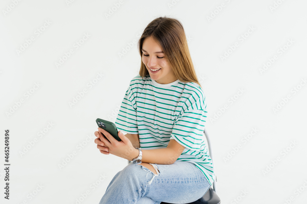 Naklejka premium Portrait of woman sitting on white chair and texting on the phone typing something with excitement on white background