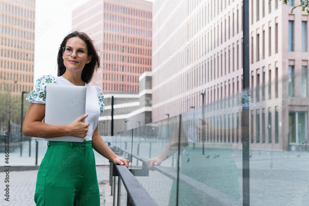 Confident businesswoman holding laptop while standing by glass railing Confident businesswoman holding laptop while standing by glass railing