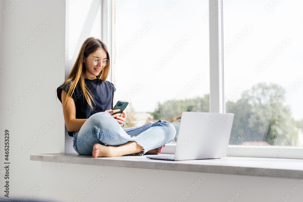 Happy young woman holds phone while sits on windowsill at home. Happy young woman holds phone while sits on windowsill at home.