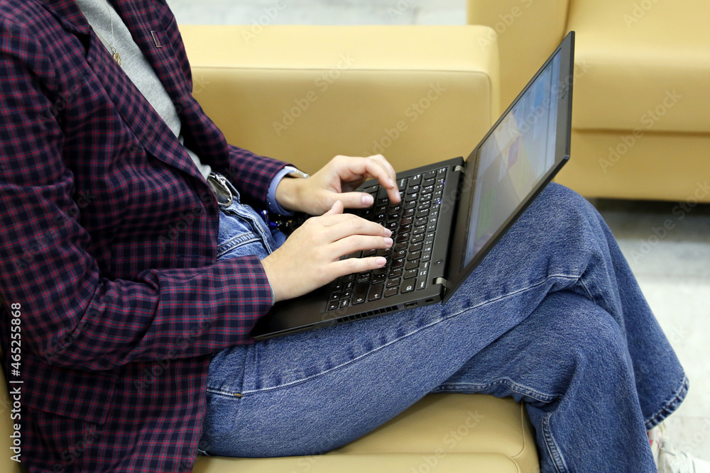 Naklejka premium Girl in jeans and checkered jacket sitting on leather sofa with laptop on her knees. Dress code, businesswoman, work in office
