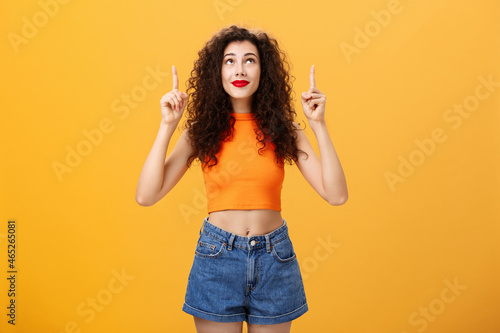 Woman wishing on shooting start believing in miracle looking and pointing hopefully up dreaming and praying in plan fullfillment standing in stylish urban cropped top over orange background photo