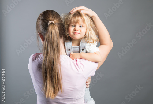 Smiling little girl looks happy in mother's arms. Beautiful baby girl, mom's hand on her head . Woman holding her daughter staying on the grey background. Rear veiw. Happy family and love concept