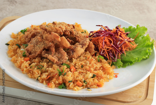 Nasi Goreng with Crispy Chicken SKin Topping, Served with Shredded Fresh Carrot and Purple Cabbage. On White Plate, Brown Table.