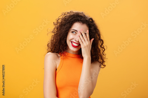 Woman being praised by coworkers feeling awkward and shy hearing congratulations with received award covering face peeking left with joyful satisfied smile posing over orange background photo