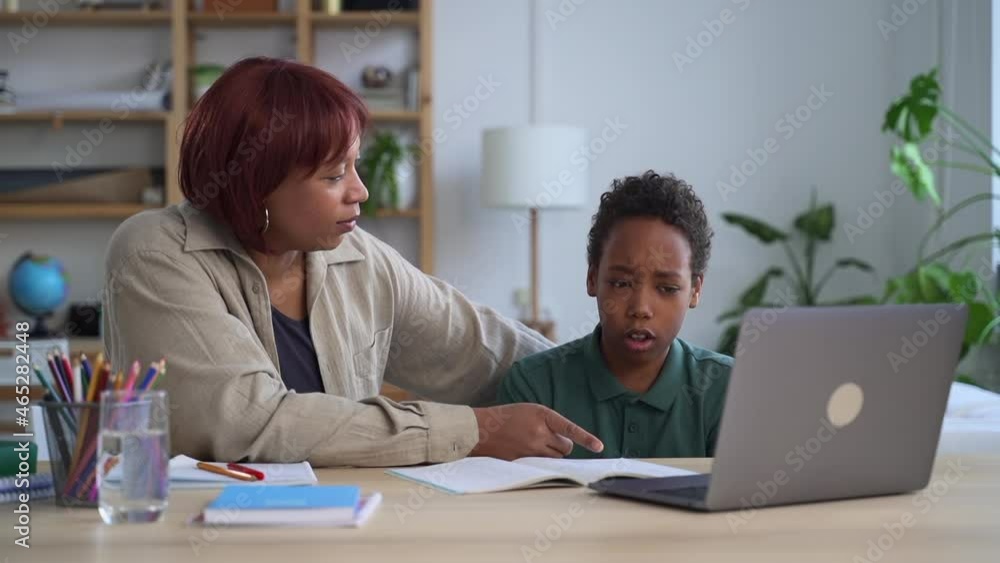 Boy writing and studying in front of laptop, mother helping him and sitting at table in home spbd. Closeup view of American African child writes in notebook and speaks something, young woman mom or Boy writing and studying in front of laptop, mother helping him and sitting at table in home spbd. Closeup view of American African child writes in notebook and speaks something, young woman mom or