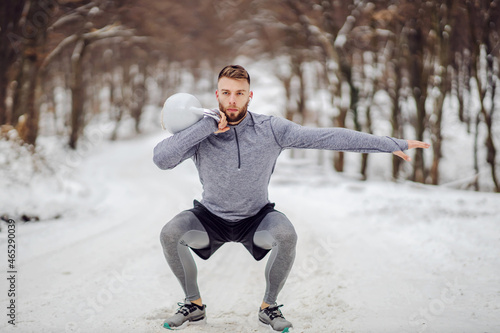 Sportsman squatting on snowy path and lifting kettlebell. Outdoor fitness, winter fitness, bodybuilding