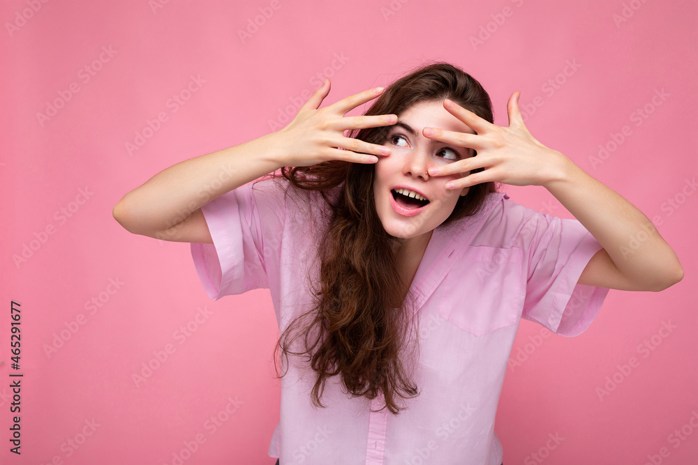 Portrait of young emotional positive surprised funny beautiful curly brunette woman with sincere emotions wearing casual pink shirt isolated over pink background with free space and covering eyes with Portrait of young emotional positive surprised funny beautiful curly brunette woman with sincere emotions wearing casual pink shirt isolated over pink background with free space and covering eyes with