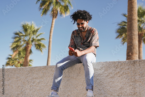 Young latin man sitting outdoor and using mobile phone with palm trees in the background photo