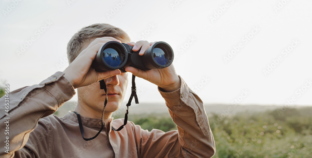 A young man looks through binoculars at a beautiful hilly landscape.The concept of hunting, travel and outdoor recreation. Banner with copy space.A traveler or hunter is observing through binoculars.