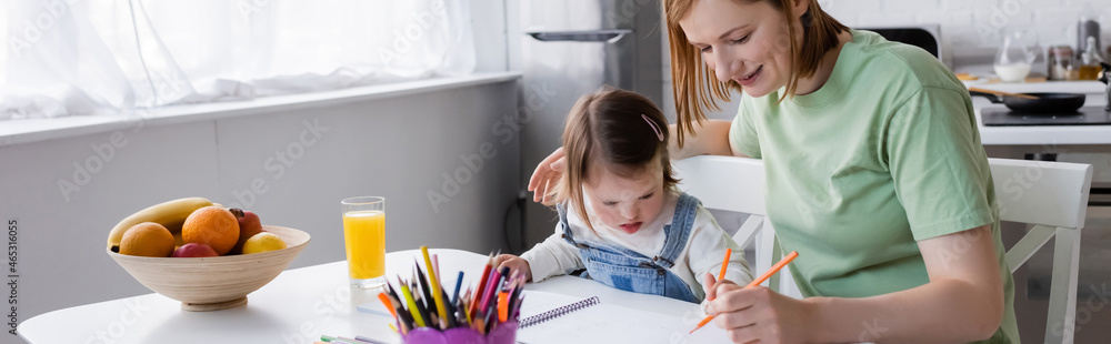 Parent and child with down syndrome drawing on paper near orange juice and fruits in kitchen, banner. Parent and child with down syndrome drawing on paper near orange juice and fruits in kitchen, banner.
