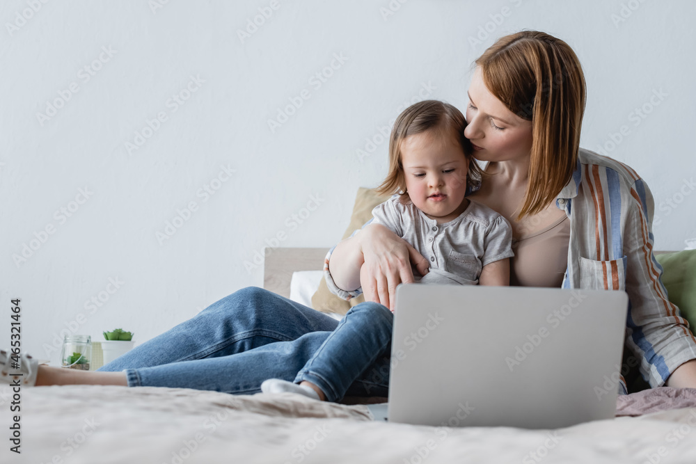 Mother kissing toddler daughter with down syndrome near laptop in bedroom. Mother kissing toddler daughter with down syndrome near laptop in bedroom.